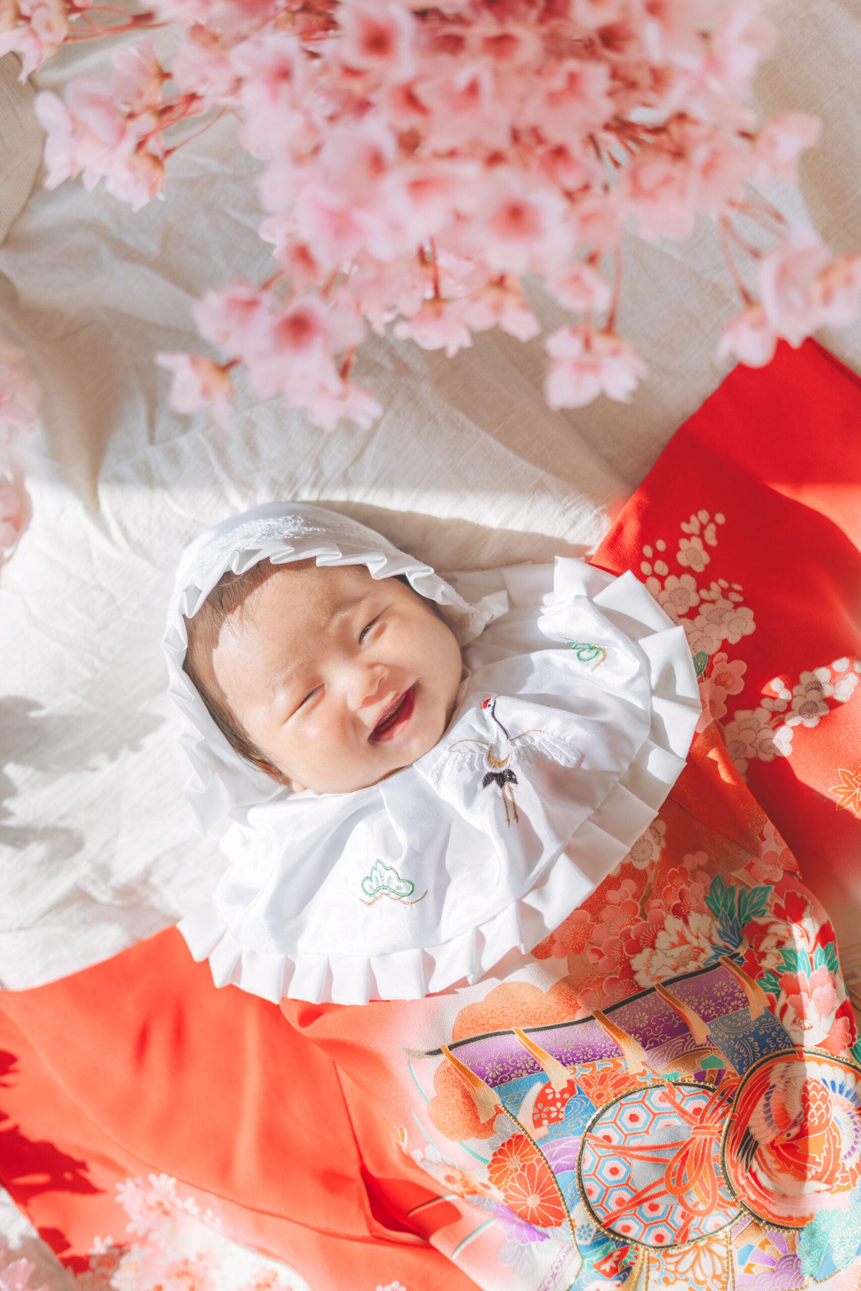 お宮参りで桜に囲まれた祝い着姿の赤ちゃんが笑顔で寝ている様子
